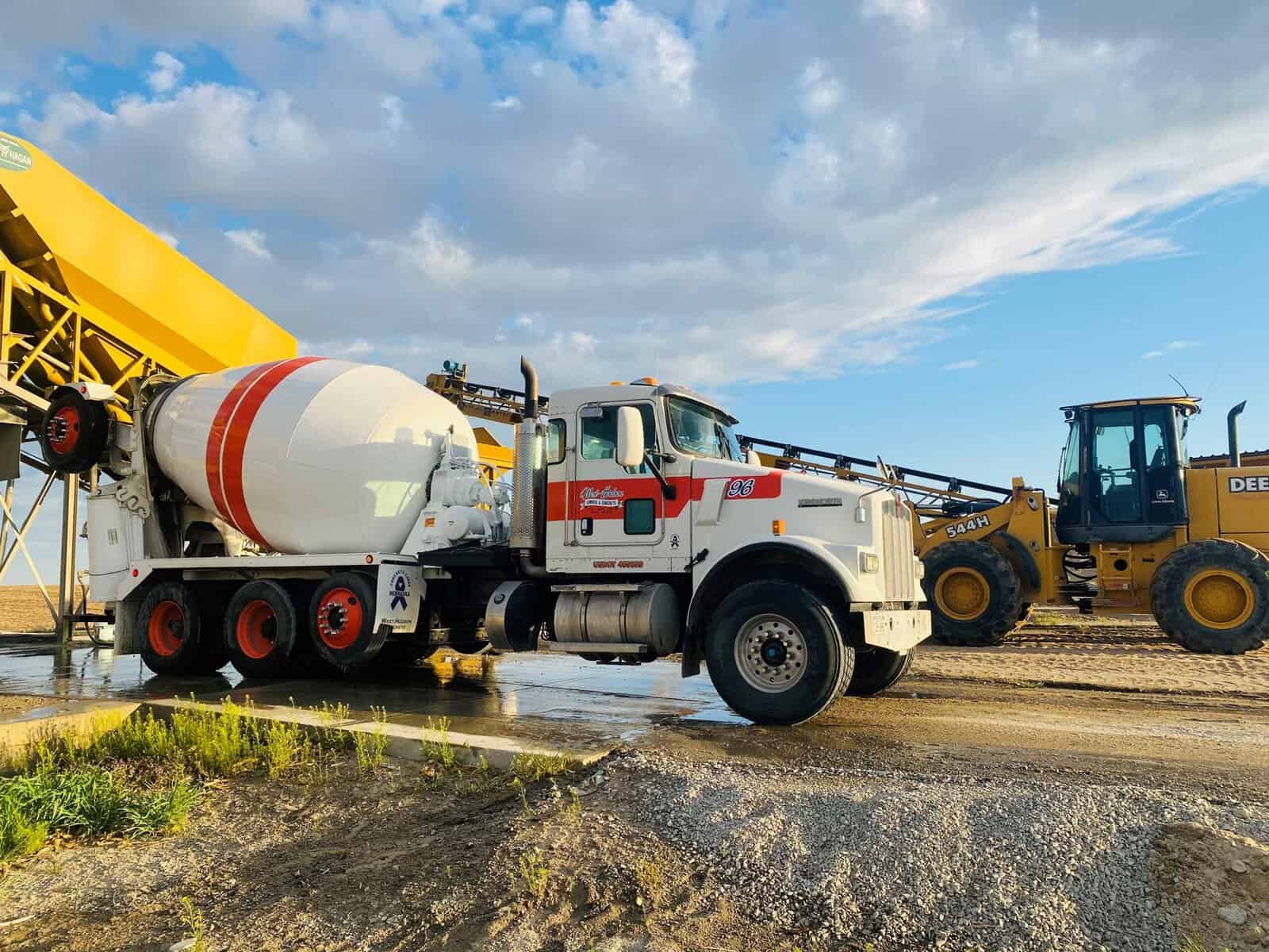 A large concrete mixer truck parked on a construction site during daytime, with construction equipment and a blue sky in the background.